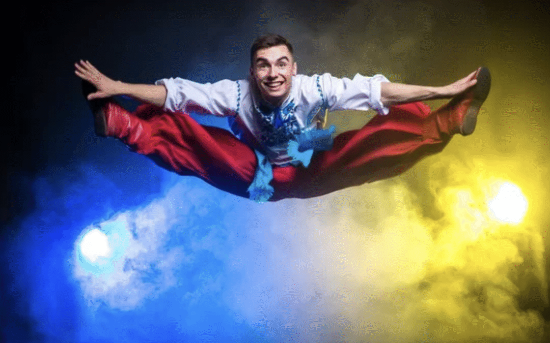 Male Cossack dancer in mid-air performing a wide straddle leap, highlighting intense groin and adductor muscle activation critical to performance.